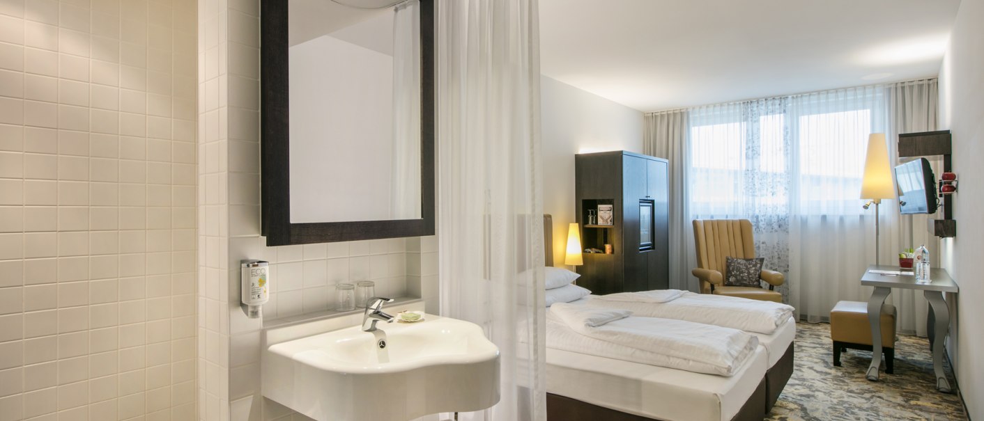 Ein modernes Hotelzimmer mit offenem Badezimmer, Doppelbett, Sessel, Schreibtisch und Fernseher. Helle Farben und stilvolle Einrichtung., © ARCOTEL Hotels