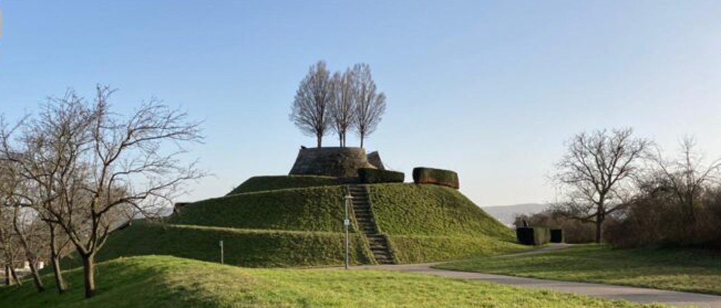Eine grasbewachsene Erhebung mit einer Treppe und Bäumen auf der Spitze, umgeben von kahlen Bäumen, unter einem klaren blauen Himmel., © SMG