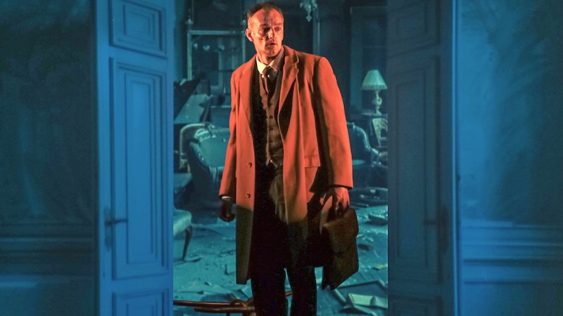 Ein Mann in einem Mantel steht in einem chaotischen Raum mit offenen Türen und einem Kronleuchter., © tri-bühne e.V. Ein Mann in einem Mantel steht in einem chaotischen Raum mit offenen Türen und einem Kronleuchter., © tri-bühne e.V.