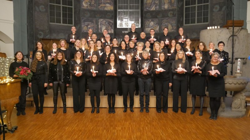 Ein Chor in schwarzer Kleidung steht in einer Kirche und hält Kerzen. Im Hintergrund sind Kirchenfenster und Dekorationen zu sehen., © Chor-Art Stuttgart e. V.