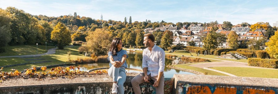 Zwei Personen sitzen auf einer bemalten Mauer im Wartbergpark. Im Hintergrund sind ein Teich, B&auml;ume und H&auml;user zu sehen. Es ist ein sonniger Tag., &copy; Stuttgart-Marketing GmbH, Sarah Schmid