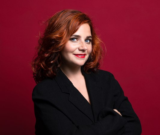 Woman with red hair and black blazer stands smiling in front of a red background., © Rosenau Kultur e.V. Woman with red hair and black blazer stands smiling in front of a red background., © Rosenau Kultur e.V.