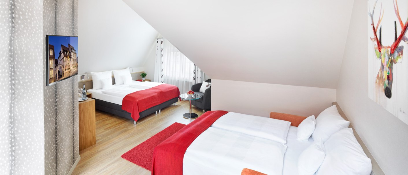 Helle Junior Suite mit zwei Betten, roten Decken, Fernseher, kleinem Tisch und buntem Hirschbild an der Wand., © Hirsch Hotel