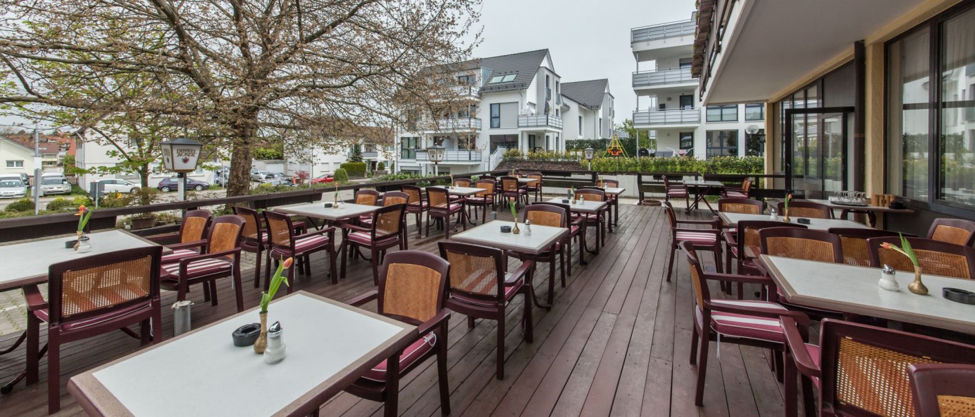 Leere Terrasse mit Tischen und Stühlen, umgeben von Wohnhäusern und einem großen Baum. Der Boden ist aus Holz., © Novum Management GmbH Leere Terrasse mit Tischen und Stühlen, umgeben von Wohnhäusern und einem großen Baum. Der Boden ist aus Holz., © Novum Management GmbH