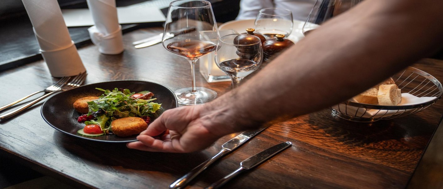 Ein Teller mit Salat und Kroketten wird auf einem gedeckten Tisch in einem Restaurant serviert. Im Hintergrund stehen Weingl&auml;ser und ein Brotkorb., &copy; Stuttgart-Marketing GmbH, Martina Denker