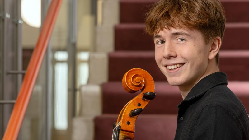 Ein junger Mann mit einem Cello l&auml;chelt auf einer Treppe. Er tr&auml;gt ein schwarzes Hemd und hat kurzes, braunes Haar., &copy; Deutsche Stiftung Musikleben