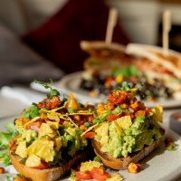Avocado toast with tomatoes and herbs, a sandwich and drinks in the background. A tea light creates a cozy atmosphere., &copy; Veganes Gold&reg;