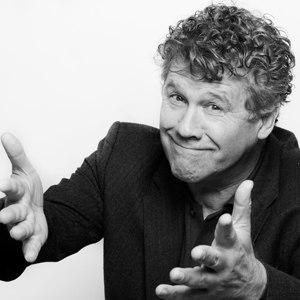 A man with curly hair is gesturing and smiling in a black-and-white photograph., © Renitenztheater Stuttgart e.V. A man with curly hair is gesturing and smiling in a black-and-white photograph., © Renitenztheater Stuttgart e.V.