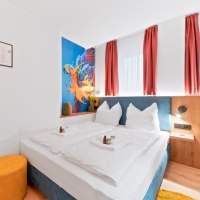 Modernes Doppelzimmer mit wei&szlig;em Bett, Papageien-Wandbild, roten Vorh&auml;ngen und gelbem Hocker. Pflegeprodukte auf dem Bett., &copy; JUNGLE N&deg;5 GmbH