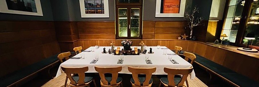 An elegant room with wooden walls, a large table with white tablecloths and chairs. Works of art hang on the walls and the window has a decorative design., © Zumbecher An elegant room with wooden walls, a large table with white tablecloths and chairs. Works of art hang on the walls and the window has a decorative design., © Zumbecher