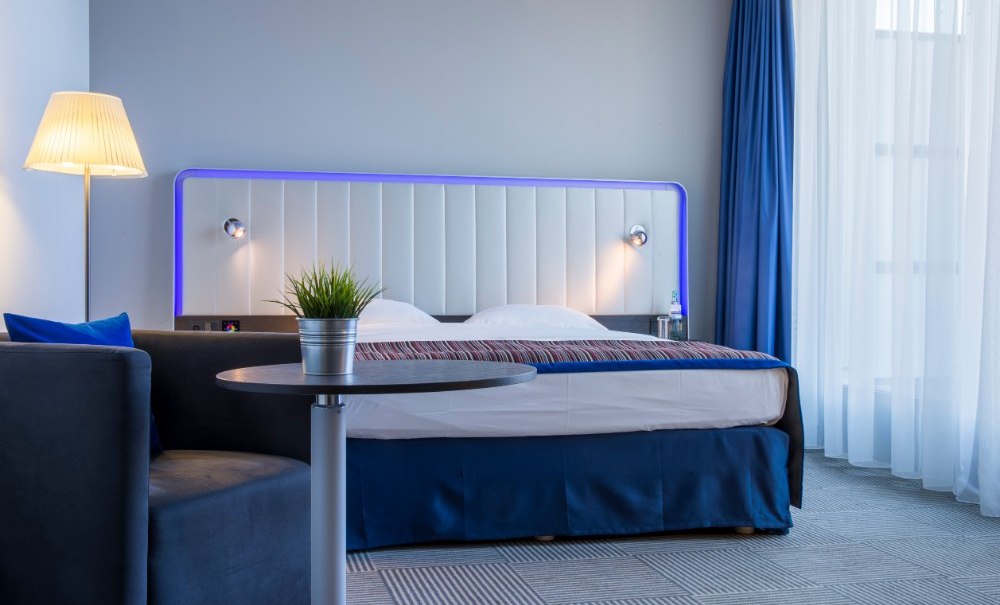 Modernes Gästezimmer mit Doppelbett, blauer Beleuchtung, Tisch mit Pflanze, Sofa und Stehlampe., © Park Inn by Radisson Stuttgart