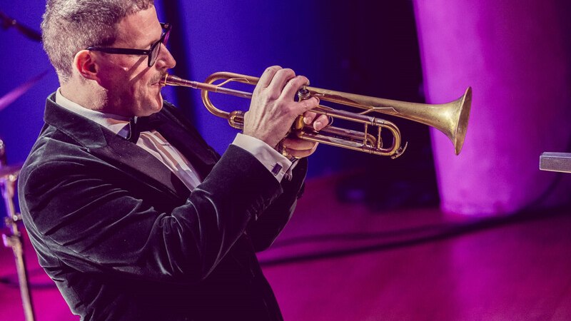 Ein Mann in Anzug spielt Trompete auf einer Bühne mit lila Beleuchtung. Er trägt eine Brille und ist in eine musikalische Darbietung vertieft., © Württembergische Staatstheater Stuttgart Ein Mann in Anzug spielt Trompete auf einer Bühne mit lila Beleuchtung. Er trägt eine Brille und ist in eine musikalische Darbietung vertieft., © Württembergische Staatstheater Stuttgart