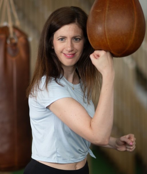 A woman in a gym poses with a punching bag. She is wearing a light blue T-shirt and smiling at the camera., &copy; Theaterhaus Stuttgart e.V.