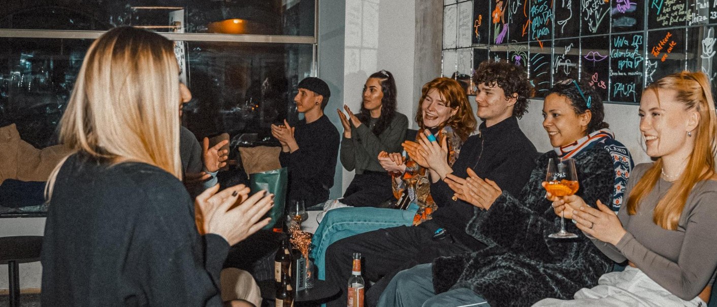 Eine Gruppe von Menschen sitzt in einem Raum, klatscht und lacht. Einige halten Getränke in der Hand. Im Hintergrund sind bunte Kritzeleien an der Wand., © Bruno, Stuttgart