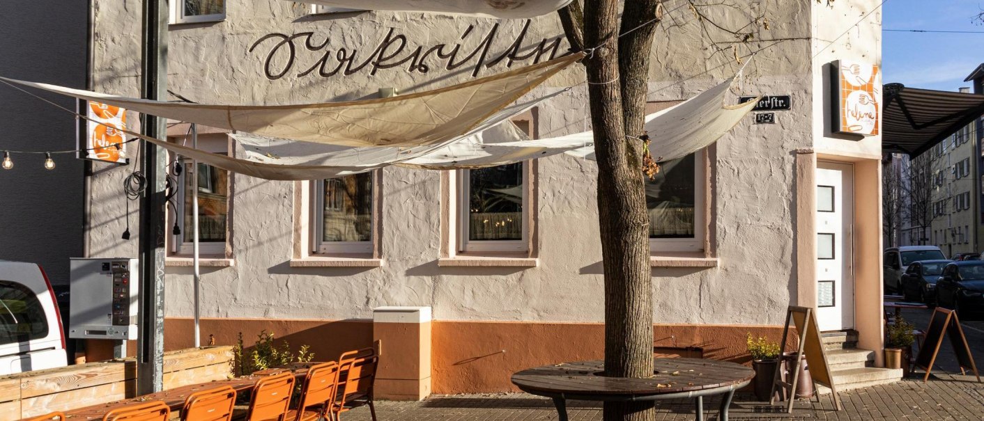 A caf&eacute; with awnings and orange chairs in the outdoor area. The fa&ccedil;ade is beige with lettering. There is a tree in front of the entrance., &copy; Stuttgart-Marketing GmbH, Sarah Schmid
