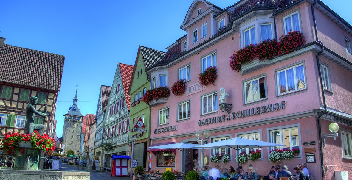 Hotel Schillerhof in einer malerischen Altstadt mit Fachwerkhäusern, einem Brunnen und Menschen, die im Freien sitzen. Sonniges Wetter und blauer Himmel., © Hotel Schillerhof