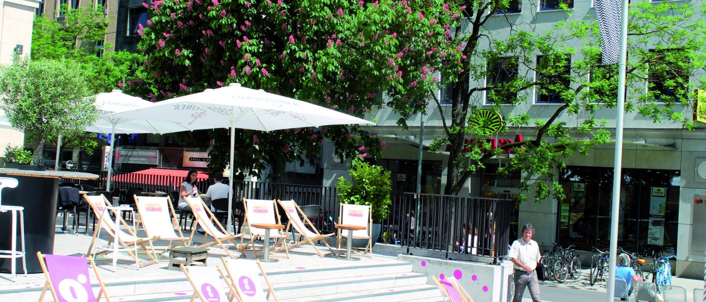 Sunny outdoor area with sun loungers and parasols in front of a building. Blossoming trees and passers-by in the background., © © Stuttgart-Marketing GmbH (SMG) Sunny outdoor area with sun loungers and parasols in front of a building. Blossoming trees and passers-by in the background., © © Stuttgart-Marketing GmbH (SMG)