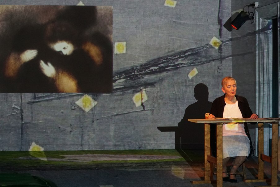 A woman sits at a table while a painting is projected in the background. The scene appears artistic and thoughtful., &copy; Katja Ritter