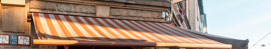 Ein Stehcafé mit gestreifter Markise und dem Schild 'Voß' an der Fassade. Die Sonne scheint auf das Gebäude., © SMG, Sarah Schmid Ein Stehcafé mit gestreifter Markise und dem Schild 'Voß' an der Fassade. Die Sonne scheint auf das Gebäude., © SMG, Sarah Schmid