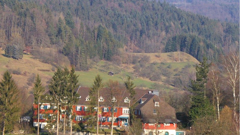 A red building with white windows stands in front of a wooded hill. Trees and bushes can be seen in the foreground., © DJH Baden-Württemberg e.V. A red building with white windows stands in front of a wooded hill. Trees and bushes can be seen in the foreground., © DJH Baden-Württemberg e.V.