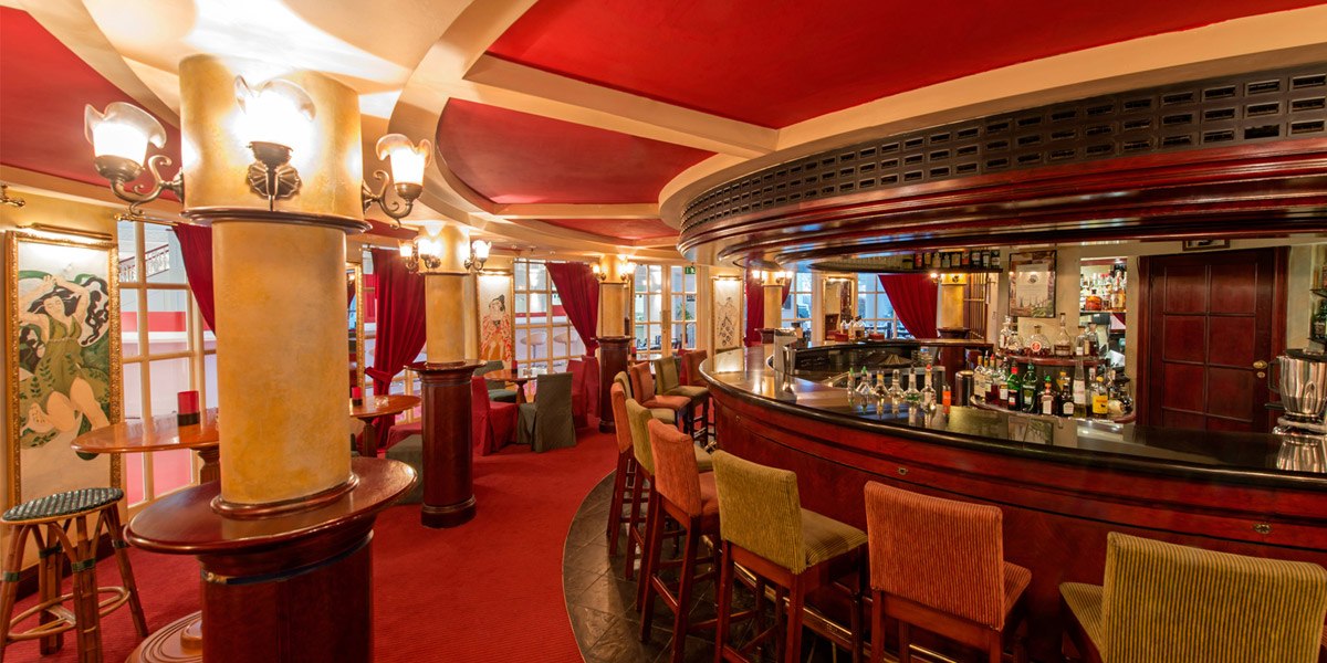 Elegante Bar mit rotem Teppich, Holzmöbeln, dekorativen Säulen und gemütlicher Beleuchtung. Sitzgelegenheiten und eine gut bestückte Theke sind sichtbar., © Dormero Hotel Stuttgart