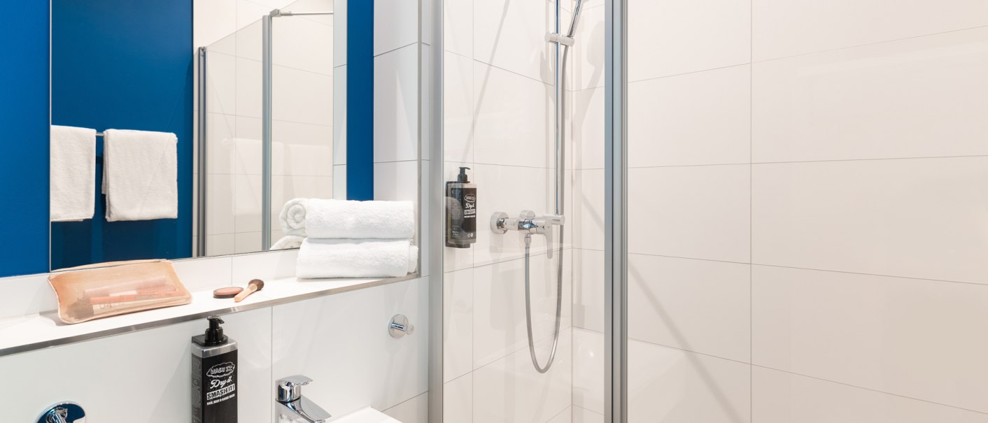 Modernes Badezimmer mit blauer Wand, Dusche, Waschbecken, Spiegel und weißen Handtüchern. Pflegeprodukte und Handtücher sind ordentlich arrangiert., © DQuadrat Living GmbH Modernes Badezimmer mit blauer Wand, Dusche, Waschbecken, Spiegel und weißen Handtüchern. Pflegeprodukte und Handtücher sind ordentlich arrangiert., © DQuadrat Living GmbH
