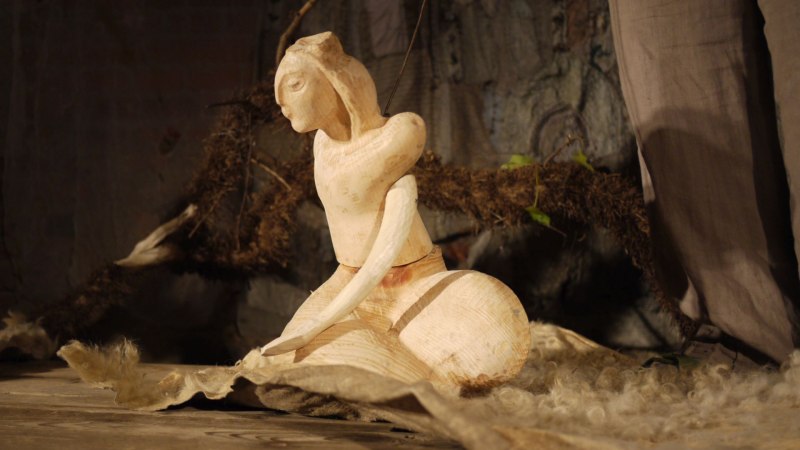 Eine Holzskulptur einer sitzenden Figur auf einem Tuch, umgeben von natürlichen Materialien in einem rustikalen Ambiente., © Theater am Faden