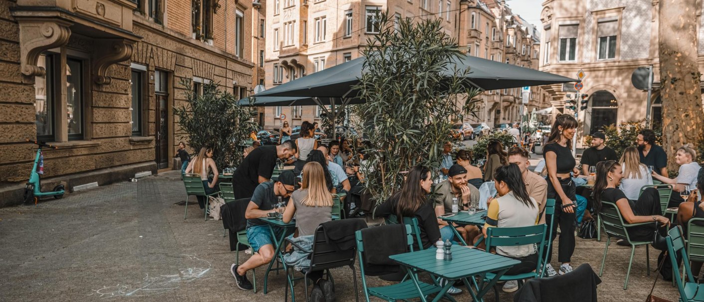 Menschen genießen das Beisammensein an grünen Tischen im Freien vor einem Café in einer städtischen Umgebung mit historischen Gebäuden., © Lupin, Stuttgart