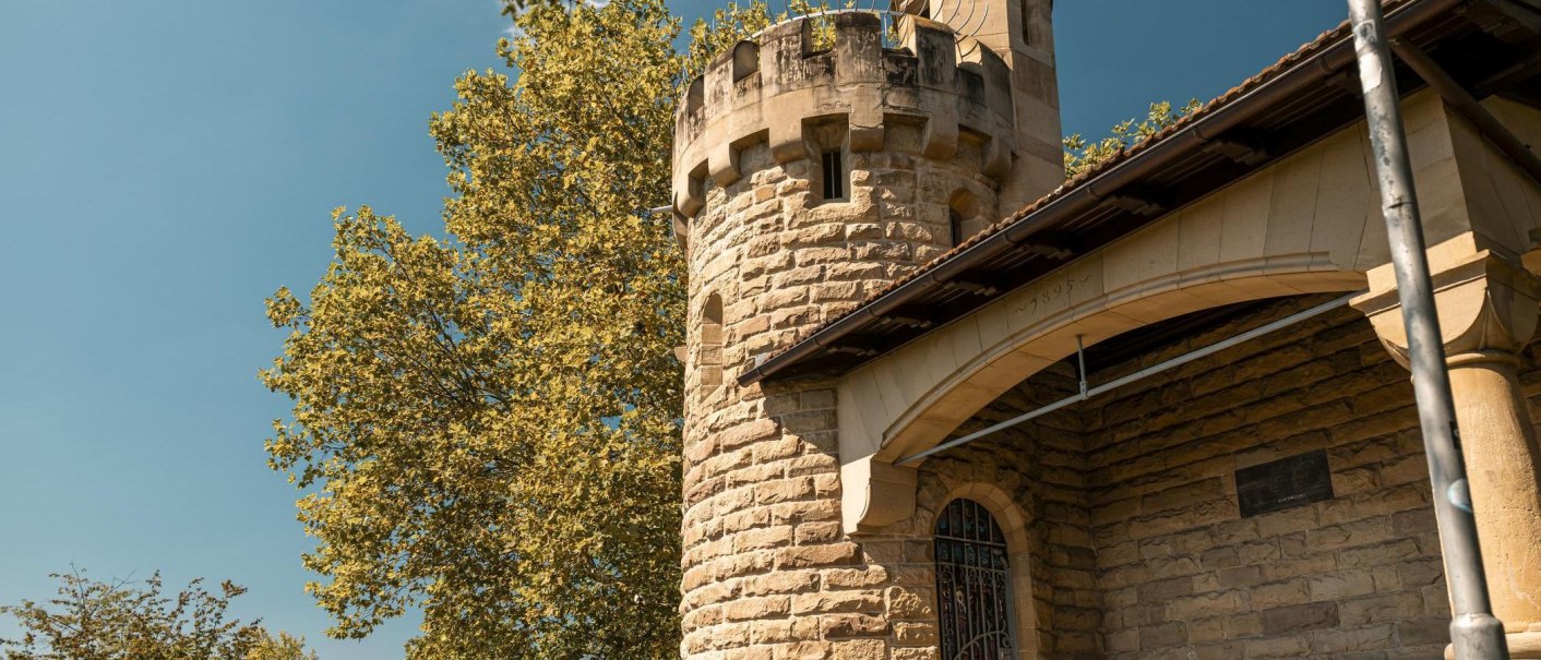 Ein steinerner Turm mit Zinnen, umgeben von Bäumen, unter einem klaren blauen Himmel. Der Turm hat ein gewölbtes Fenster und ein Dach aus Ziegeln., © Stuttgart Marketing GmbH, Sarah Schmid