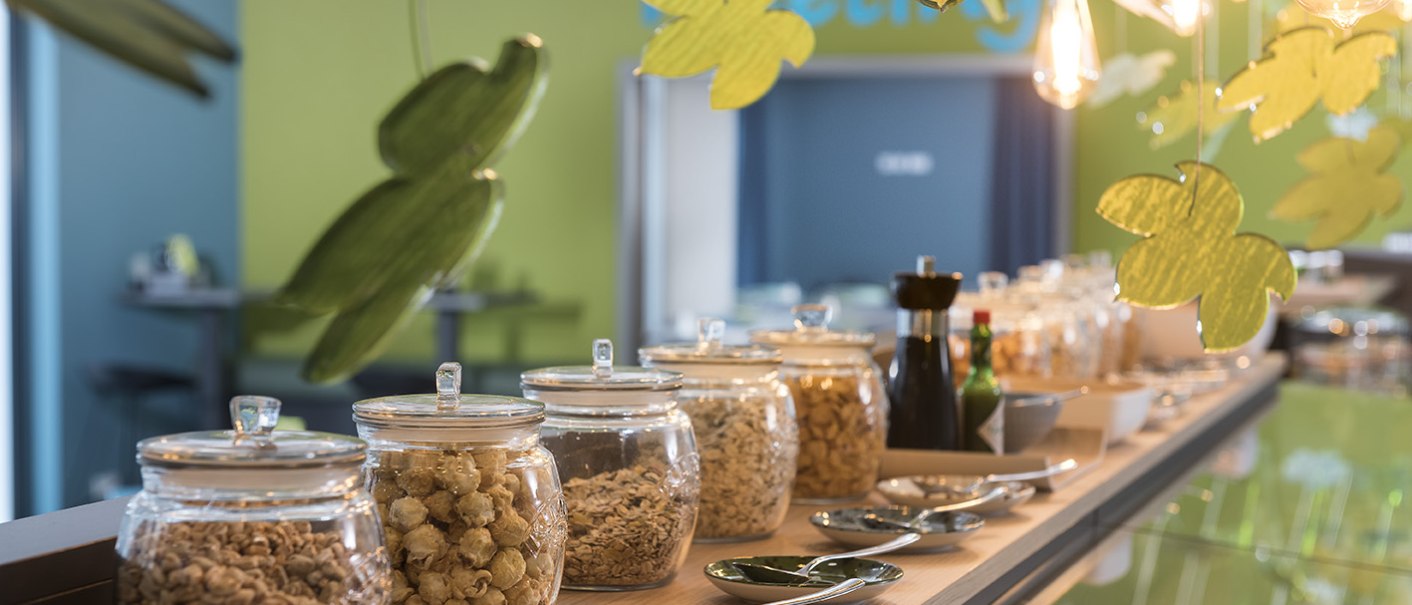 Ein Frühstücksbuffet mit Gläsern voller Cerealien, dekoriert mit hängenden Blättern und warmem Licht., © ibis Styles Stuttgart Vaihingen