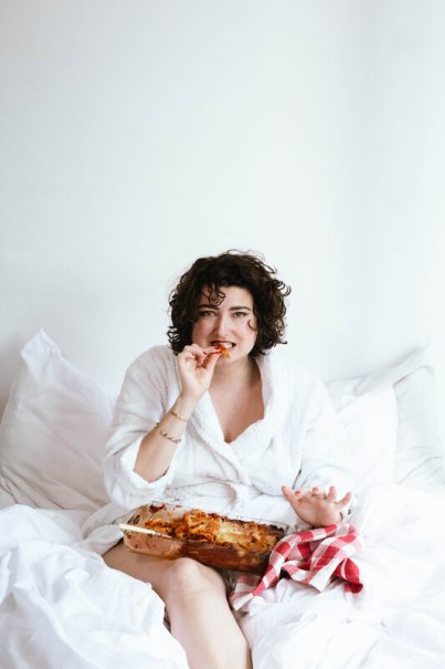 A person in a white bathrobe sits in bed and eats lasagna from a casserole dish. She is holding a piece of lasagna in her hand., © Rosenau Kultur e.V. A person in a white bathrobe sits in bed and eats lasagna from a casserole dish. She is holding a piece of lasagna in her hand., © Rosenau Kultur e.V.