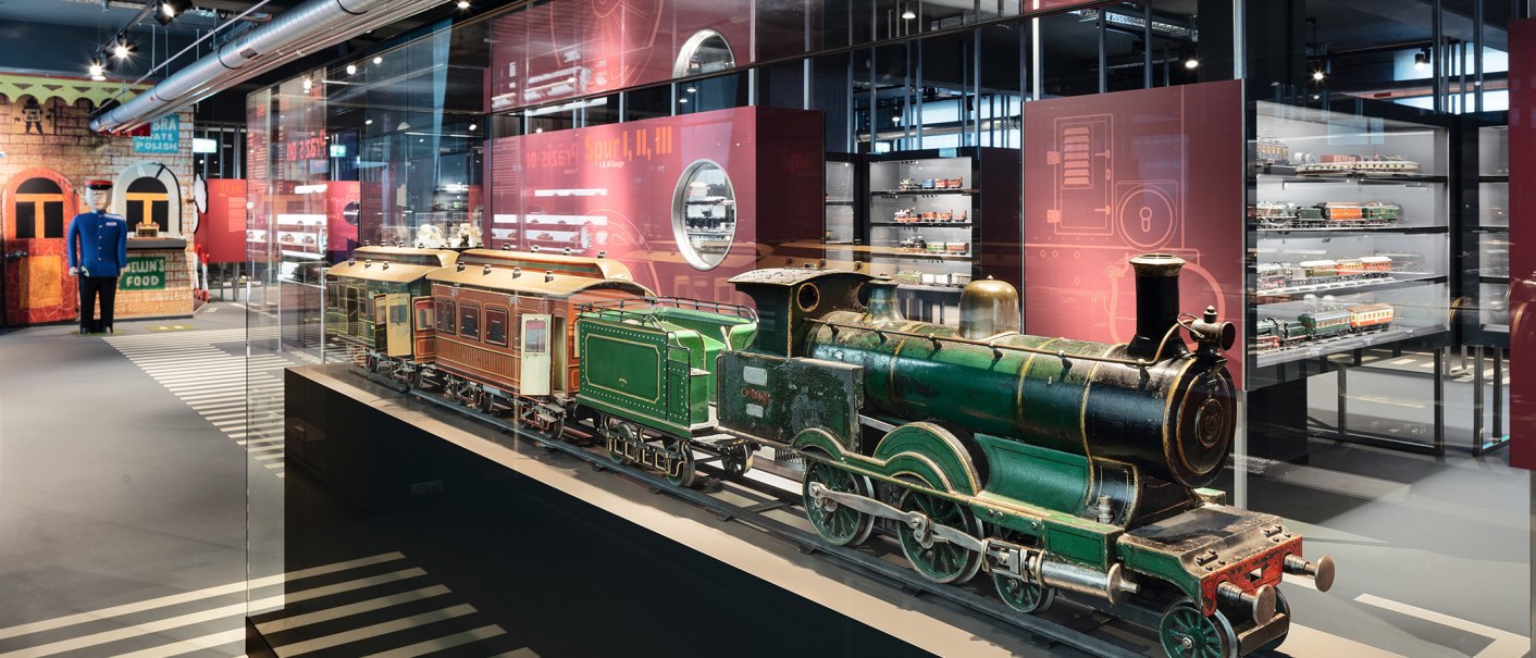A green model locomotive in a glass display case, surrounded by other model trains in a museum exhibition room., © Christian Höhn A green model locomotive in a glass display case, surrounded by other model trains in a museum exhibition room., © Christian Höhn