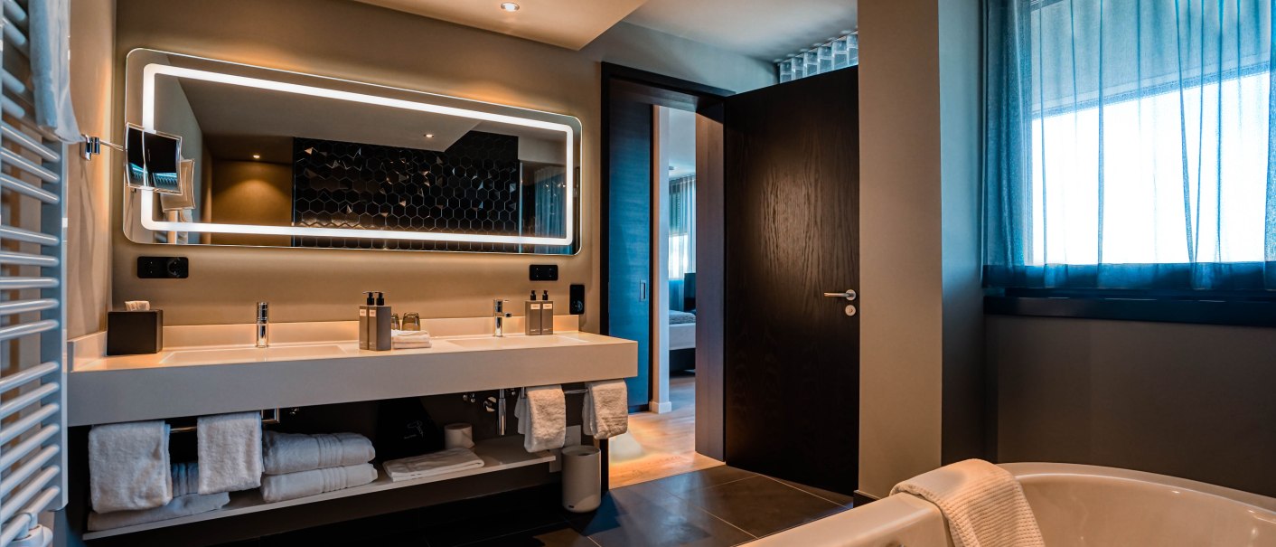 Modernes Badezimmer mit Doppelwaschbecken, beleuchtetem Spiegel und Badewanne. Durch die Tür sieht man ein Schlafzimmer. Elegantes Design mit dunklen Fliesen., © CLOUD N°7 APARTMENTS GmbH Modernes Badezimmer mit Doppelwaschbecken, beleuchtetem Spiegel und Badewanne. Durch die Tür sieht man ein Schlafzimmer. Elegantes Design mit dunklen Fliesen., © CLOUD N°7 APARTMENTS GmbH