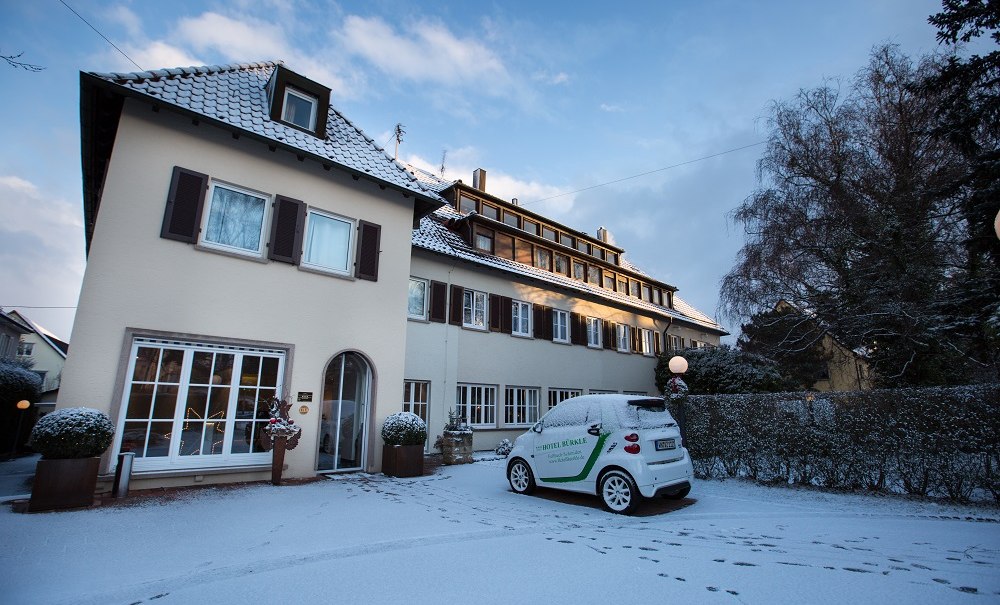 A hotel building in winter, covered in snow. A car with the word "Hotel" on it is parked in front of it. The sky is blue with a few clouds., &copy; Hotel B&uuml;rkle