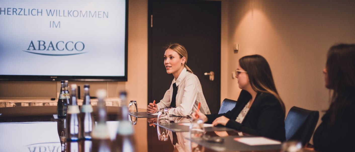 Drei Personen sitzen in einem Konferenzraum an einem Tisch. Im Hintergrund ist ein Bildschirm mit der Aufschrift 'Herzlich Willkommen im Abacco' zu sehen., © Abacco Hotels GmbH Drei Personen sitzen in einem Konferenzraum an einem Tisch. Im Hintergrund ist ein Bildschirm mit der Aufschrift 'Herzlich Willkommen im Abacco' zu sehen., © Abacco Hotels GmbH