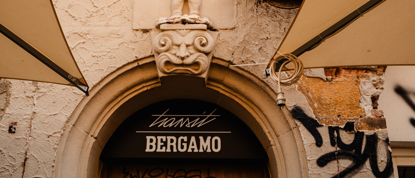 Steinrelief eines Kindes über einem Eingang mit der Aufschrift 'transit Bergamo'. Der Putz ist bröckelig, Graffiti und Markisen sind sichtbar., © SMG_Sarah Schmid