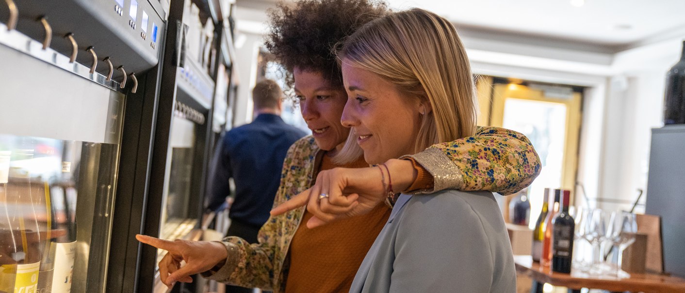Zwei Frauen stehen vor einem Weinspender und zeigen auf verschiedene Weinflaschen. Im Hintergrund sind weitere Flaschen und Gläser zu sehen., © Stuttgart Marketing GmbH, Fotografin Martina Denker