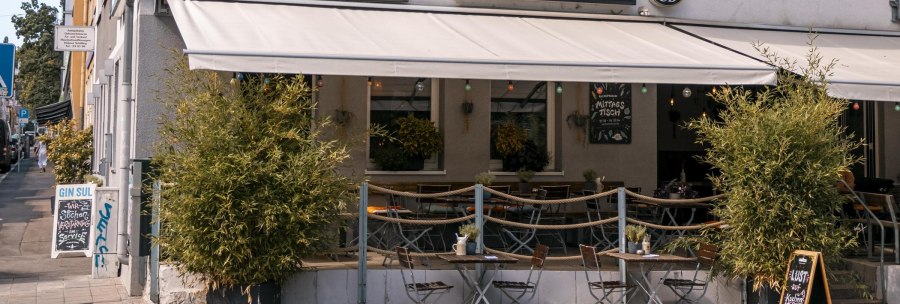 Café 'Sutsche' mit Außenbereich, Tischen und Stühlen auf dem Gehweg, umgeben von Pflanzen., © SMG, Sarah Schmid