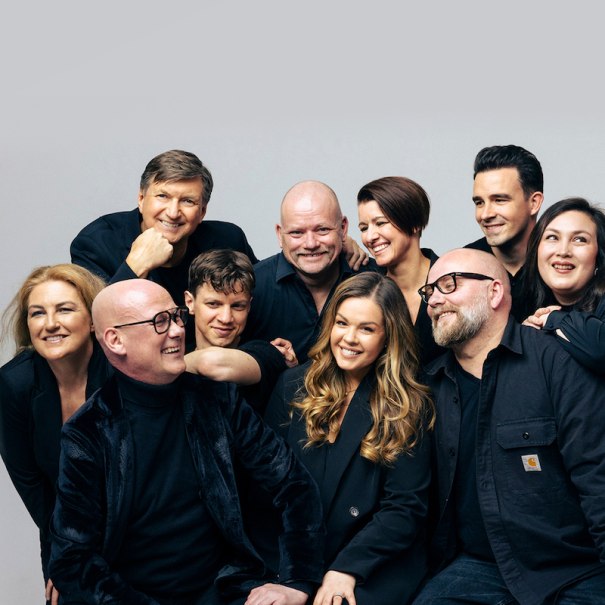 Zehn Personen in schwarzer Kleidung l&auml;cheln fr&ouml;hlich in die Kamera. Sie stehen eng beieinander vor einem neutralen Hintergrund., &copy; GCM Konzerte