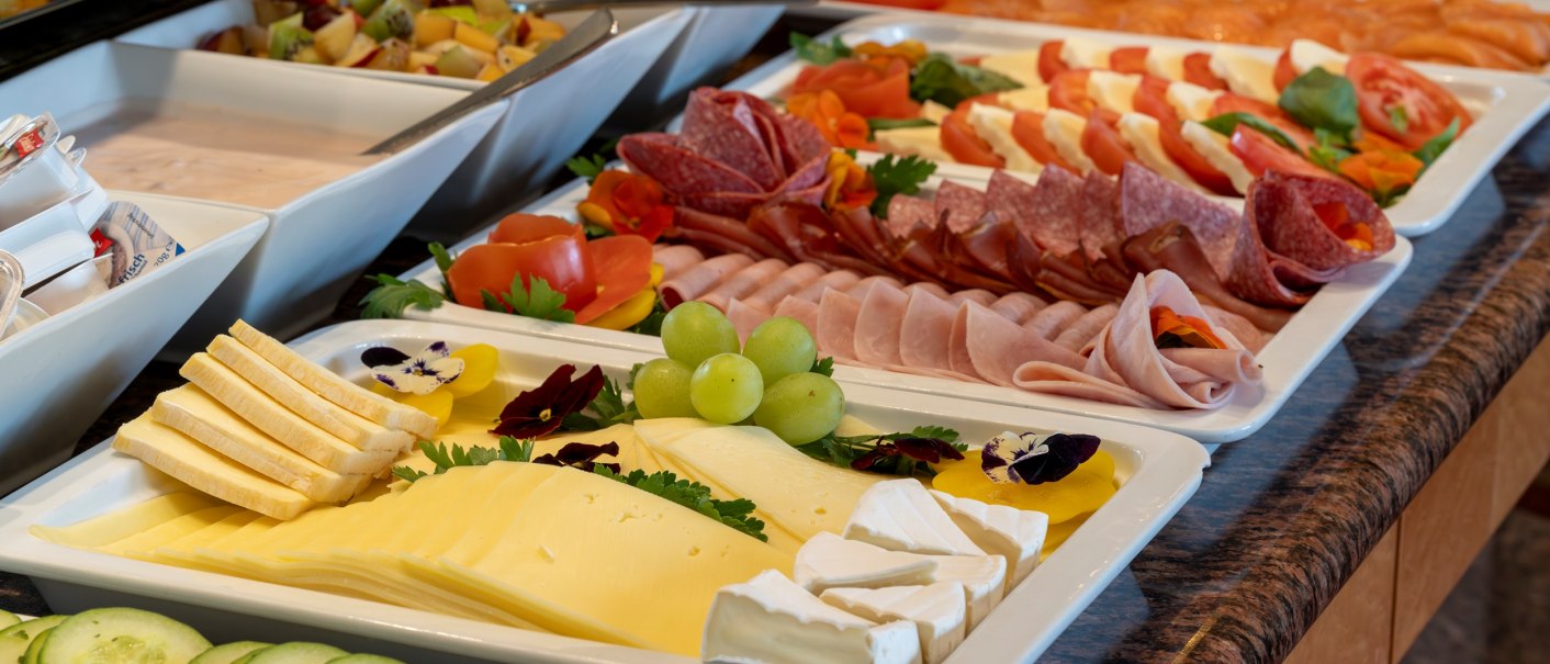 Ein reichhaltiges Frühstücksbuffet mit verschiedenen Käsesorten, Wurst, frischem Obst und Gemüse auf weißen Platten angerichtet., © PLAZA INN Rieker Stuttgart Hauptbahnhof Ein reichhaltiges Frühstücksbuffet mit verschiedenen Käsesorten, Wurst, frischem Obst und Gemüse auf weißen Platten angerichtet., © PLAZA INN Rieker Stuttgart Hauptbahnhof