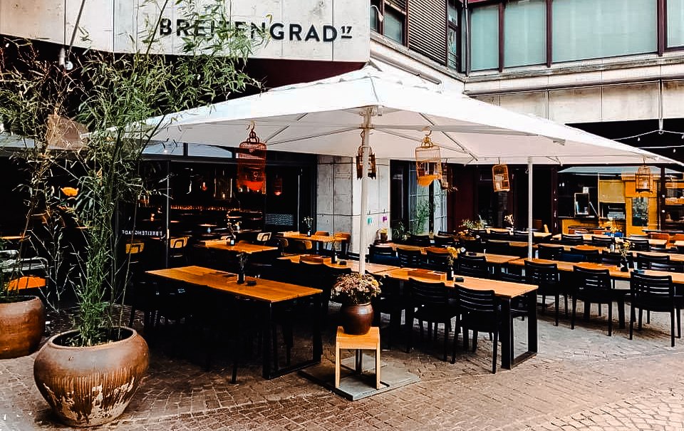 Außenbereich eines Restaurants mit Holztischen und Stühlen unter einem großen Sonnenschirm. Pflanzen in Töpfen dekorieren den Bereich., © Breitengrad17 Stuttgart