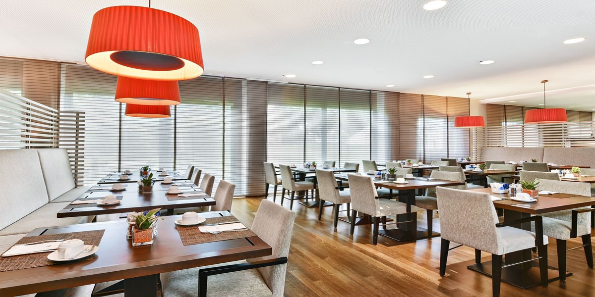 A modern breakfast room with set tables, red pendant lights, and large windows with blinds., © NH Ludwigsburg A modern breakfast room with set tables, red pendant lights, and large windows with blinds., © NH Ludwigsburg
