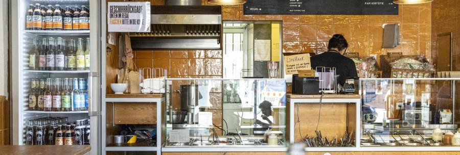 A snack bar with drinks fridge, counter and menu board. One person works behind the counter. The walls are tiled in orange., © SMG, Sarah Schmid