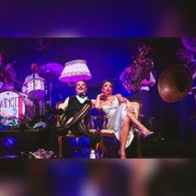 Eine Band auf der Bühne mit zwei Personen im Vordergrund, die auf Stühlen sitzen. Im Hintergrund sind Instrumente und eine Lampe sichtbar., © links im Bild