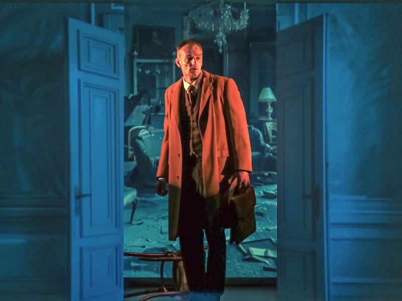 Ein Mann in einem Mantel steht in einem chaotischen Raum mit offenen Türen und einem Kronleuchter., © tri-bühne e.V. Ein Mann in einem Mantel steht in einem chaotischen Raum mit offenen Türen und einem Kronleuchter., © tri-bühne e.V.