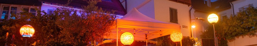 An illuminated half-timbered house with a white tent in the garden. Colorful lanterns and fairy lights create a cozy atmosphere at night., © Weingut Ruoff
