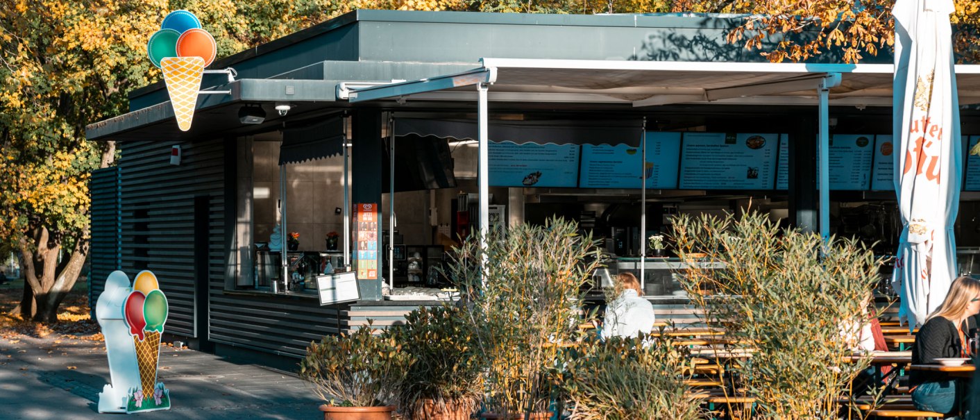 Ein Biergarten mit Eisdiele, umgeben von herbstlichen B&auml;umen. Menschen sitzen an Holztischen, w&auml;hrend ein Eis-Schild den Eingang ziert., &copy; SMG, Sarah Schmid
