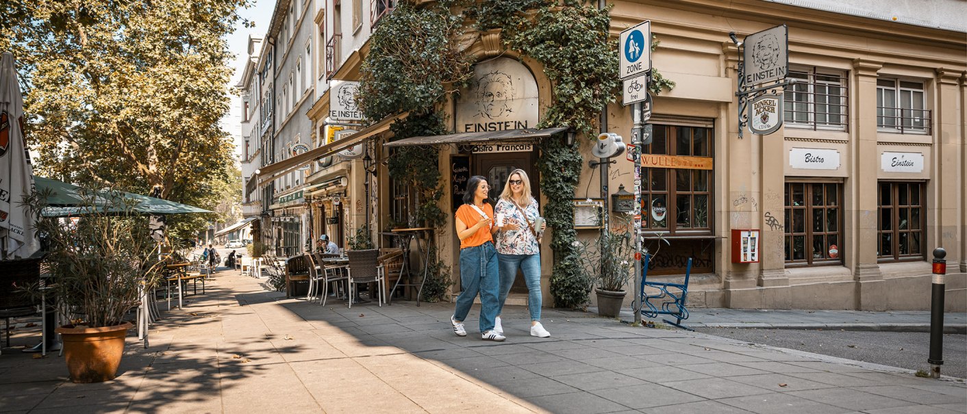 Zwei Frauen spazieren lächelnd am Café Einstein vorbei. Die Straße ist gesäumt von Bäumen und Außensitzplätzen. Es herrscht eine entspannte Atmosphäre., © Stuttgart-Marketing GmbH, Sarah Schmid Zwei Frauen spazieren lächelnd am Café Einstein vorbei. Die Straße ist gesäumt von Bäumen und Außensitzplätzen. Es herrscht eine entspannte Atmosphäre., © Stuttgart-Marketing GmbH, Sarah Schmid