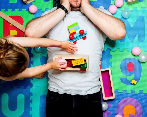 Mann liegt überrascht auf bunter Spielmatte, umgeben von Bauklötzen und Spielzeug. Ein Kind reicht ihm Spielzeug. Humorvolle Szene., © Theaterhaus Stuttgart e.V. Mann liegt überrascht auf bunter Spielmatte, umgeben von Bauklötzen und Spielzeug. Ein Kind reicht ihm Spielzeug. Humorvolle Szene., © Theaterhaus Stuttgart e.V.