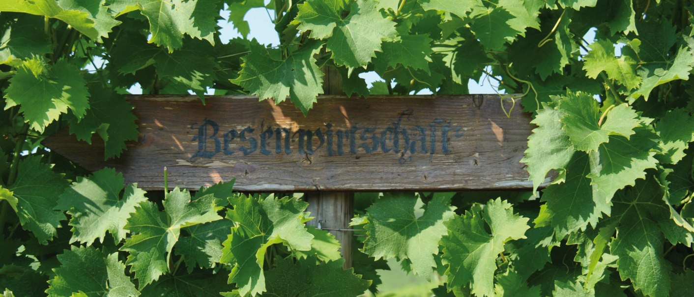 Ein Holzschild mit der Aufschrift 'Besenwirtschaft' ist von grünen Weinblättern umgeben., © SMG Ein Holzschild mit der Aufschrift 'Besenwirtschaft' ist von grünen Weinblättern umgeben., © SMG